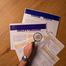 A person holding a magnifying glass over a piece of paper with an escape room puzzle game scenario titled 'The Uncle's Treasure'. Other scenarios like 'Coffee with a Pirate' and 'The Manor' are also visible nearby.